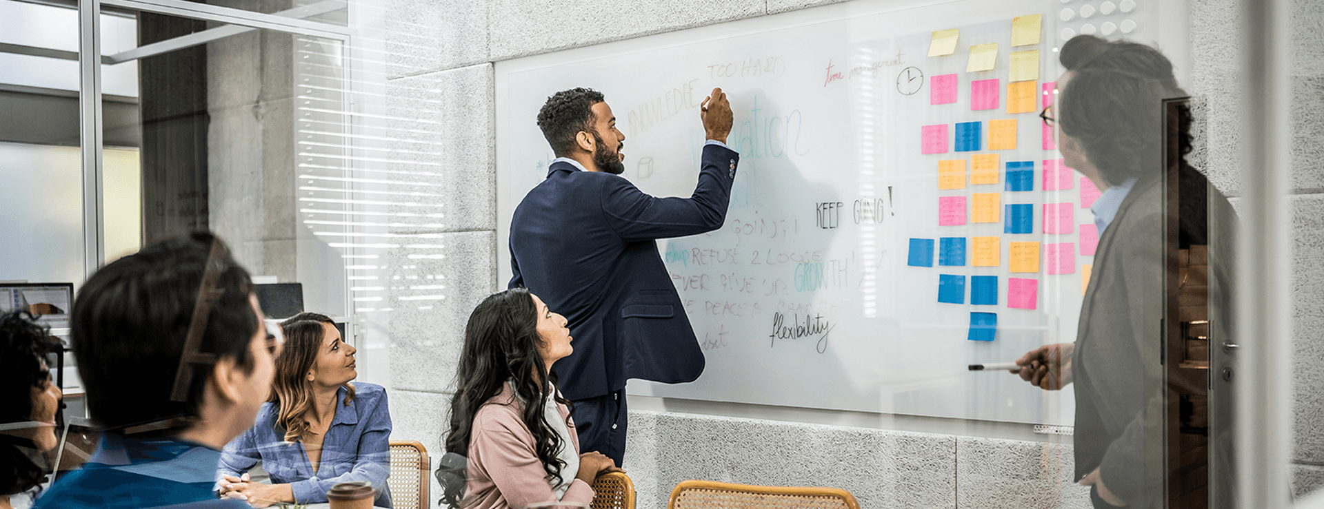 Ein Geschäftsmann schreibt in einem Anzug mit einem Marker auf eine große, transparente Whiteboard-Wand in einem modernen Büro, während eine Gruppe von Kollegen bei einem Meeting zuschaut.