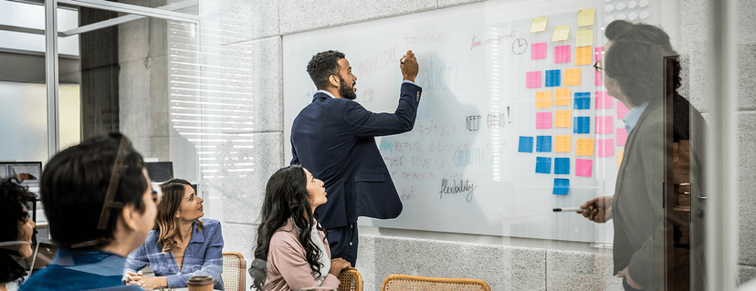 Ein Geschäftsmann schreibt in einem Anzug mit einem Marker auf eine große, transparente Whiteboard-Wand in einem modernen Büro, während eine Gruppe von Kollegen bei einem Meeting zuschaut.