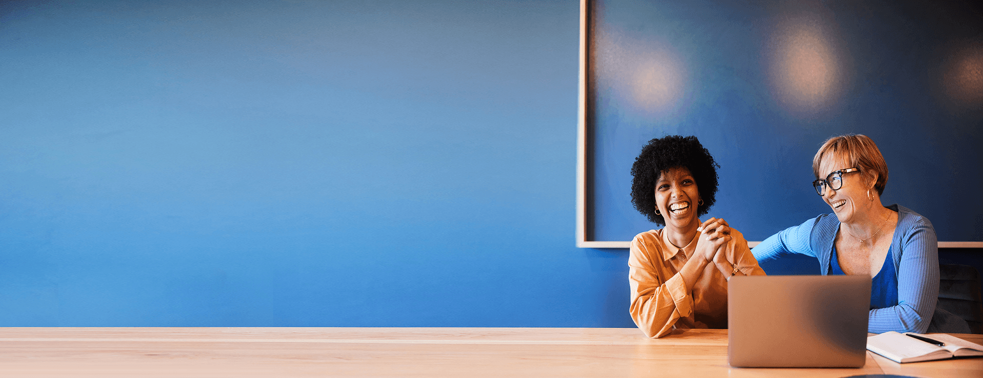 Zwei lachende Frauen, eine junge und eine ältere, sitzen an einem Holztisch vor einer leuchtend blauen Wand mit einem Laptop und Notizbuch.