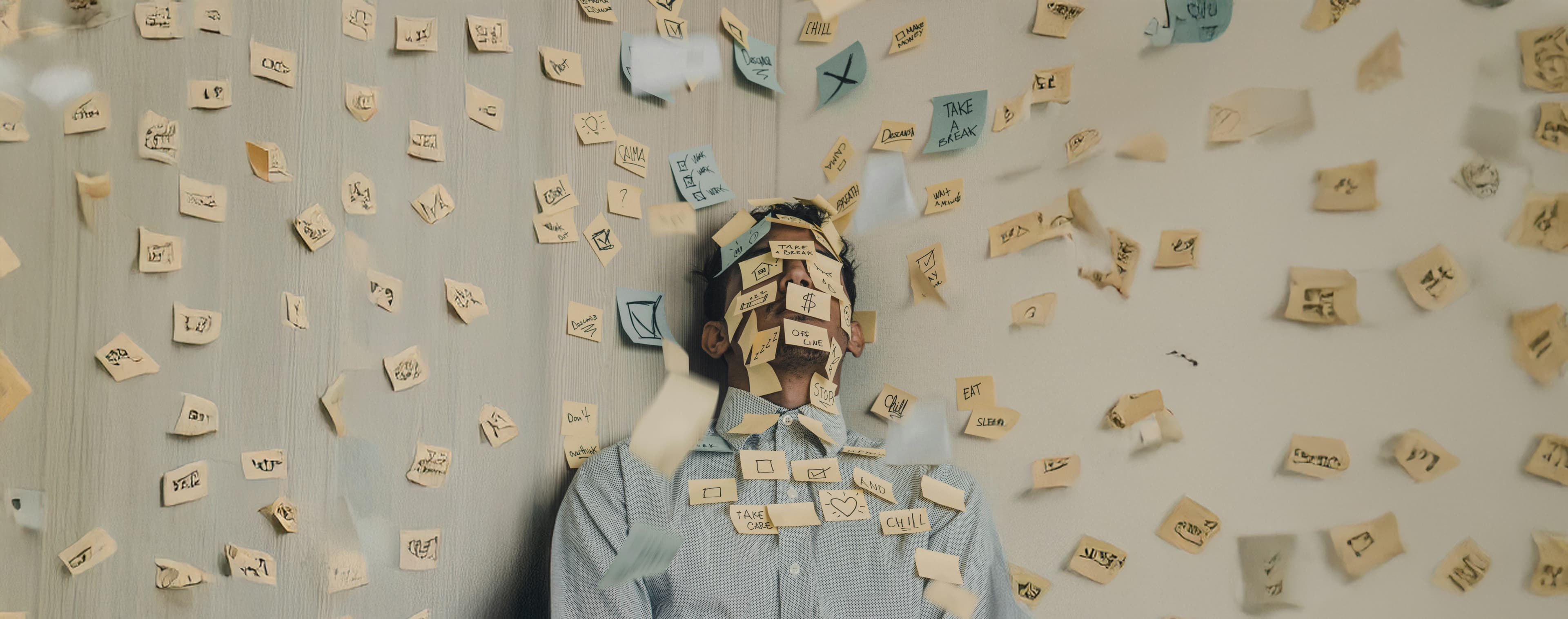 Ein Typ sitzt total überfordert da, umgeben von Haftnotizen. Hunderte davon kleben an der Wand hinter ihm und schweben um ihn herum in der Luft. Auf den Notizen stehen Kritzeleien und Erinnerungen wie „Mach mal Pause“, „Entspann dich“ und „Iss was“, die das Gefühl von Burnout und mentaler Überlastung zeigen.