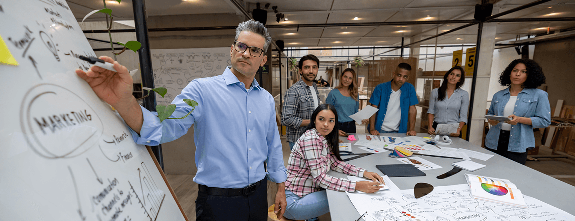 Mittelalter Mann mit Brille in blauem Hemd schreibt MARKETING an ein Whiteboard, während sein diverses Team im modernen Büro zusieht.